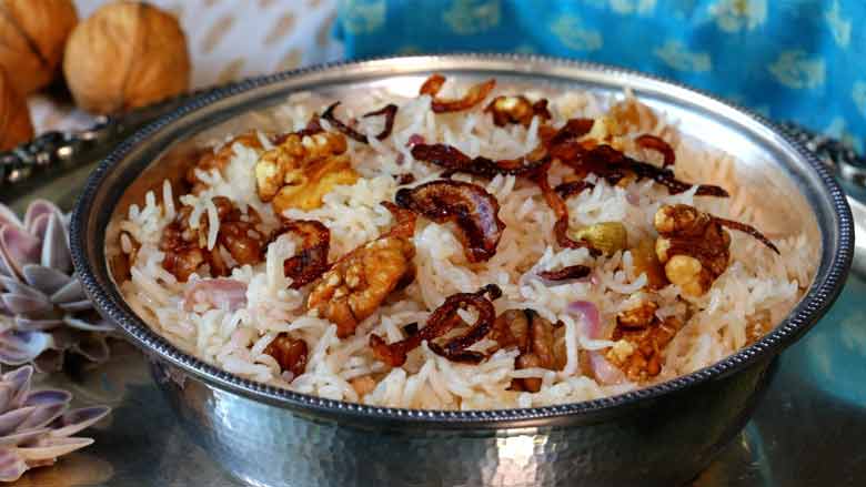 The ghee rice is served in a traditional silver bowl on a silver platter with shell flowers in the platter. There is a blue printed cloth and whole and split California walnuts in the background - Kerala Ghee Rice with California Walnuts Recipe